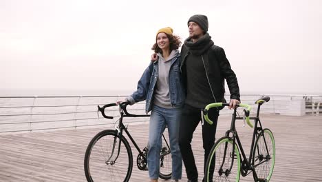 Moderna-Pareja-Joven-Hipster-Caminando-Junto-Con-Sus-Bicicletas-Cerca-Del-Mar-En-El-Día-De-Otoño-Abrazando