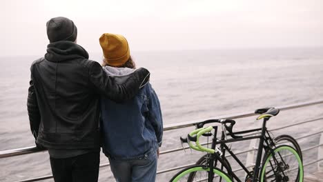 Back-Portrait-Of-Young-Couple-Standing-Near-The-Sea,-Love-Couple-Concept,-Couple-With-Bikes,-Couple-Wear-Warm-Clothes,-Guy-With-A-Girl-Standing-On-Sea-Wooden-Pier