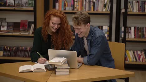 Front-View-Of-Two-Young-Students-Female-With-Long-Red-Hair-Dressed-In-Black-Sweater-And-Male-In-Blue-Shirt-Talking-In-The-Library