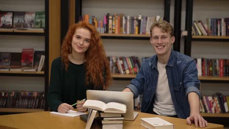 Dos-Jóvenes-Estudiantes-Europeos-Masculinos-Y-Femeninos-Sentados-En-La-Mesa-Con-Libros-Y-Laptop-En-La-Biblioteca-Preparándose-Para-Los-Exámenes