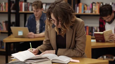 Estudiante-Europea-Concentrada-Preparándose-Para-El-Examen-Y-Escribiendo-Notas-Mientras-Se-Sienta-En-La-Mesa-En-La-Biblioteca-Universitaria-Con-Sus-Compañeros-De-Clase-Estudiando-Libros,-Trabajando-En-Laptopn-En-Segundo-Plano