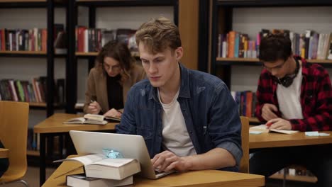 Blonde-Guy-Sitting-In-College-Library-And-Working-On-Laptop,-Preparing-For-Exams-Then-Looks-On-A-Camera-And-Smiling