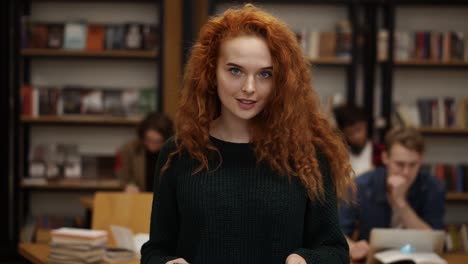 Portrait-Of-An-Attractive-Long-Red-Haired-European-Girl-Student-Standing-In-High-School-Library-Smiling-Looking-At-Camera-And-Smiling