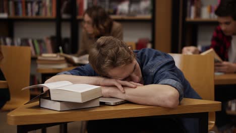 Young-European-Student-In-A-College-Library-Or-Class-Sleeping-On-Desk-With-Pile-Of-Books-On-It