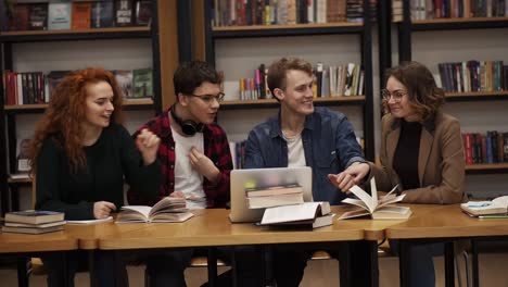 Group-Of-Young-Students-Talking-Using-Laptop-Explaining-Course-Work-Study-Together-In-Library