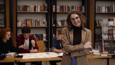 Portrait-Of-An-Attractive-Short-Haired-European-Girl-Student-In-Glasses-And-Brown-Jacket-Standing-In-High-School-Library-Smiling-Looking-At-Camera-And-Smiling