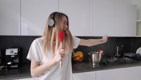 Young-Woman-In-Headphones-Dancing-And-Pretending-Singing-In-Kitchen-At-Home