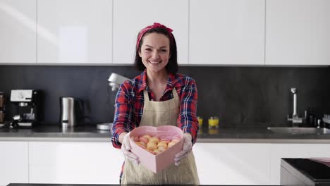 Smiling-Woman-In-Apron-Presents-Box-With-Macaroons-Cookies-At-The-Kitchen