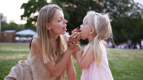 Portrait-Of-Mother-And-A-Small-Daughter,-Spends-Time-Together-In-A-City-Park-On-A-Picnic-They-Bite-The-Little-Cake-From-Sides-Together