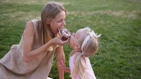 Portrait-Of-Mother-And-A-Small-Daughter,-Spends-Time-Together-In-A-City-Park-On-A-Picnic-They-Bite-A-Tasty-Donut-From-Sides-Together