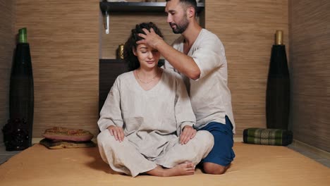 Attractive-Thai-Massagist-Is-Massaging-Young-Woman's-Head-Standing-Behind-Her