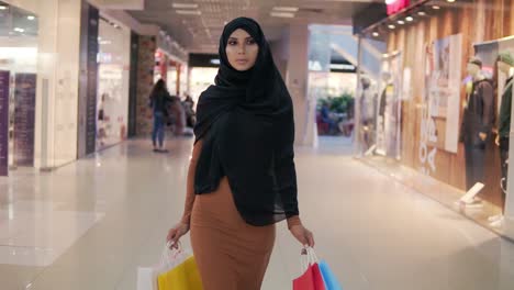 Porträt-Einer-Stilvollen-Jungen-Muslimischen-Frau-In-Schwarzem-Hijab-Und-Engem-Braunem-Kleid-Mit-Taschen-In-Ihren-Händen-Nach-Dem-Einkaufen,-Das-Durch-Ein-Einkaufszentrum-Geht