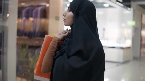 Wunderschöne-Junge-Muslimische-Frau-In-Schwarzem-Hijab-Mit-Verschiedenen-Taschen-Auf-Der-Schulter,-Die-Am-Einkaufszentrum-Vorbeigehen