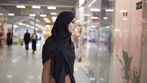 Schöne-Muslimische-Frau-Im-Hijab,-Die-Durch-Ein-Großes-Einkaufszentrum-Geht-Und-Durch-Fenster-Nach-Verkäufen-Sucht