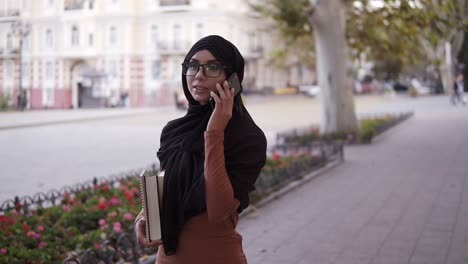 A-Young-Muslim-Woman-In-A-Traditional-Headscarf-Chatting-With-Friends-On-A-Smartphone-On-The-Street-While-Carrying-Her-College-Books