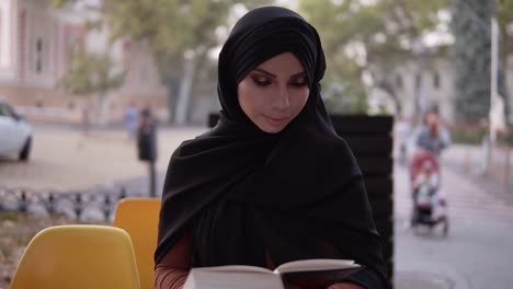 Chica-Con-Hiyab-Negro-Leyendo-Un-Libro-Mientras-Se-Sienta-En-Un-Café-Al-Aire-Libre,-Preparándose-Para-El-Examen,-Educación-Entre-Mujeres-Musulmanas