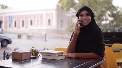 Joven-Y-Bonita-Mujer-Musulmana-Con-Hiyab-Hablando-Por-Teléfono-Y-Sonriendo,-Sentada-En-Un-Café,-Alegre