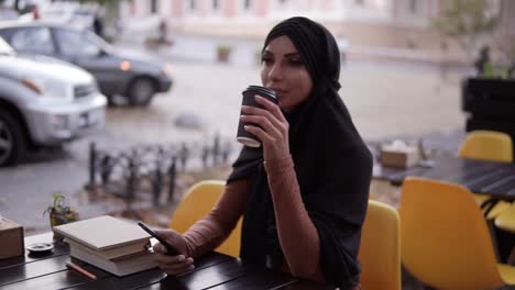 Schöne-Junge-Muslimische-Frau,-Die-Auf-Dem-Smartphone-Chattet-Und-Am-Wochenende-In-Einem-Café-Kaffee-Aus-Der-Kartontasse-Trinkt