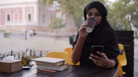 Schöne-Junge-Moslemische-Frau,-Die-Auf-Dem-Smartphone-Chattet-Und-Am-Wochenende-In-Einem-Café-Kaffee-Aus-Der-Kartontasse-1-Trinkt