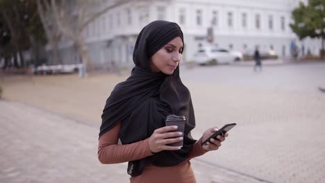 Attractive-Young-Woman-In-Hijab-With-A-Perfect-Make-Up-Walking,-Drinking-Coffee-And-Looking-At-Her-Cell-Phone-Screen-And-Smiling-To-The-Camera