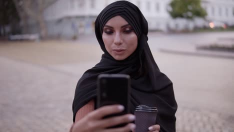 Young-Woman-In-Black-Hijab-With-A-Perfect-Make-Up-Walking,-Drinking-Coffee