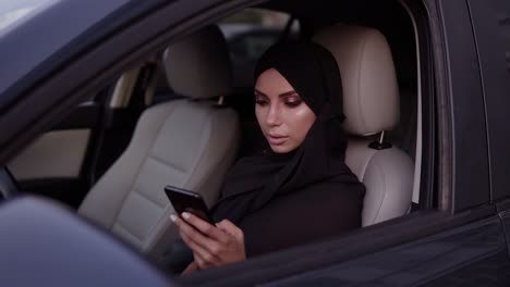 Attractive-Young-Woman-In-Black-Hijab-Sitting-In-Her-Car,-Thoughtfully-Looking-At-Her-Smartphone-Screen