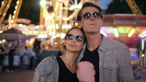 Portrait-Of-Stylish-Hipster-Young-Couple-In-Sunglasses-Are-Standing-And-Looking-Thoughtfully-For-Attractions-At-Amusement-Park-At-Night