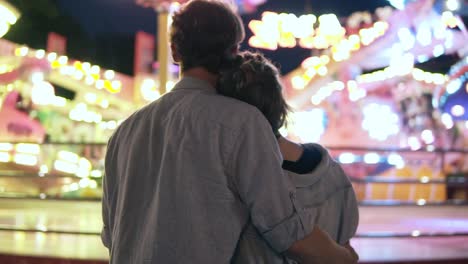 Young-Couple-Visiting-An-Amusement-Park-Arcade-Together-And-Hugging-While-Standing-Next-To-A-Carousel-Ride-With-Lights-At-Night