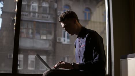 Attractive-Sexy-Young-Guy-Sitting-Near-Panoramic-Window-Typing-Something-On-His-Laptop