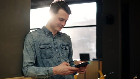 Young-Attractive-Sexy-Guy-In-Denim-Shirt-Standing-And-Scrolling-Something-On-His-Smartphone
