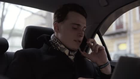 Attractive-Young-Guy-Is-Holding-Laptop-On-His-Laps-While-Talking-To-Someone-On-The-Phone,-Sitting-And-Riding-In-A-Car