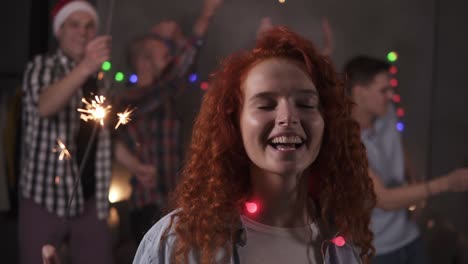 Precious-Curly-Redhaired-Girl-Posing-For-Camera-Dancing-And-Smiling,-Waving-Her-Bengal-Light-While-Her-Friends-Dancing-On-The-Blurred-Background-In-Decorated-Room