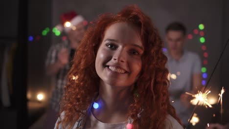 Cheerful-Curly-Redhaired-Girl-With-Colorful-Lights-On-Neck-Posing-For-Camera-Smiling,-Holding-Her-Bengal-Light-While-Her-Friends-Dancing-On-The-Blurred-Background-In-Decorated-Room