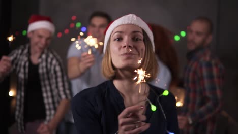 Pretty-Girl-With-Colorful-Lights-On-Neck-And-Santa-Hat-Posing-For-Camera-Smiling,-Waving-Her-Bengal-Light-While-Her-Friends-Dancing-On-The-Blurred-Background-In-Decorated-Room