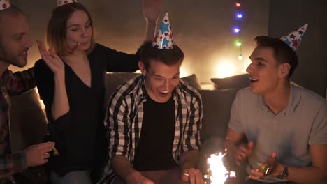 Friends-In-Colorful-Caps-Celebrating-Birthday-Party,-Surprising-Friend-With-Cake-With-Burning-Sparkling-Stick