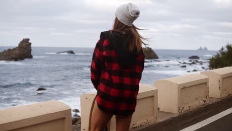 Stylish,-Sporty-Builded-Woman-In-Hat,-Long-Plaid-Coat-And-White-Sneakers-Enjoying-Time-By-The-Seaside-On-A-Cloudy-Day-At-Sunset,-Walking,-Holding-A-Skateboard,-Watching-Stormy-Ocean-From-Promenade,-Slow-Motion