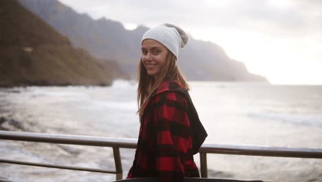 Close-Up-Footage-Sporty-Woman-In-Hat,-Plaid-Coat-Enjoying-Time-By-The-Seaside-On-A-Cloudy-Day-At-Sunset,-Walking-Freerly,-Holding-A-Skateboard