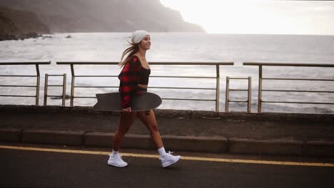 Side-View-Of-Muscular,-Sporty-Woman-In-Hat,-Coat-And-Shorts-Enjoying-Time-By-The-Seaside-On-A-Cloudy-Day-At-Sunset,-Walking-Freerly,-Holding-A-Skateboard-1