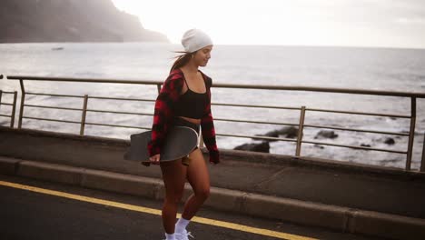 Cheerful,-Sporty-Woman-In-Hat,-Coat-And-Shorts-Enjoying-Time-By-The-Seaside-On-A-Cloudy-Day-At-Sunset,-Walking-Freerly,-Holding-A-Skateboard