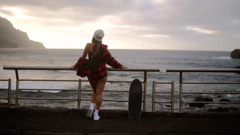 Full-Length-Sporty-Builded-Woman-In-Hat,-Plaid-Coat-And-White-Sneakers-Enjoying-Time-By-The-Seaside-On-A-Cloudy-Day-At-Sunset,-Standing-Leaning-On-Railings-With-Skateboard,-Watching-Stormy-Ocean-From-Promenade