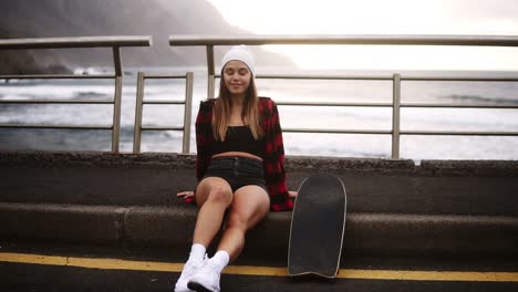 Smiling-Girl-Sitting-With-Longboard-At-The-Border-At-Coastline-Asphalt-Road-On-Background-Hills-And-Foggy-Ocean