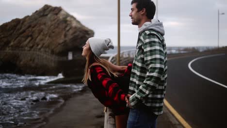 Woman-And-Man-Gazes-At-Ocean-Young-Couple-Shown-From-The-Side-Look-Out-Over-An-Ocean-In-Front-Of-The-Hills,-Talking,-Embracing