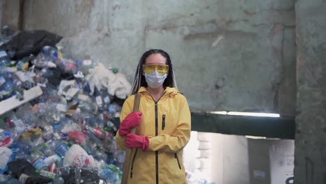 Portrait-Of-A-Girl-With-Dreadlocks,-In-Yellow-Jacket-And-Mask,-Holding-Large-Duck-Shovel,-Standing-At-A-Plastic-Recycling-Factory