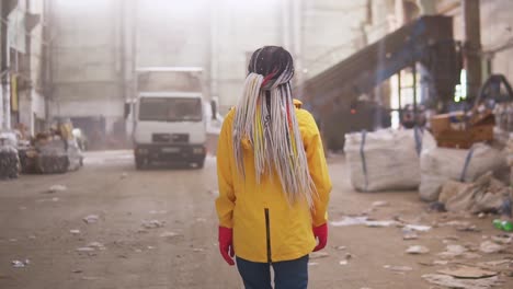 Tracking-Rare-View-Footage-Of-A-Girl-In-Protective-Working-Clothes-Walking-By-Waste-Recycling-Factory,-Huge-Area-With-Used-Bottles,-Different-Waste-And-White-Truck-On-Background