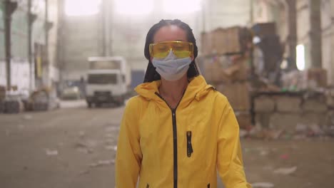 A-Positive-Girl-In-Protective-Working-Clothes-Yellow-Jacket,-Eyeglasses-And-Mask-Walking-By-Recycling-Factory-And-Smiling