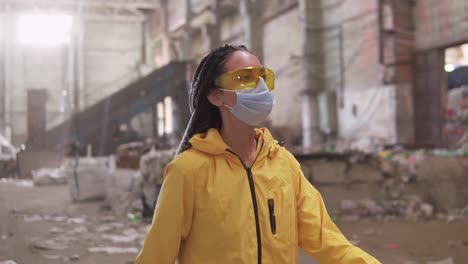 A-Confident-Girl-With-Dreadlocks-In-Protective-Working-Clothes-Yellow-Jacket,-Eyeglasses-And-Mask-Walking-By-Recycling-Factory