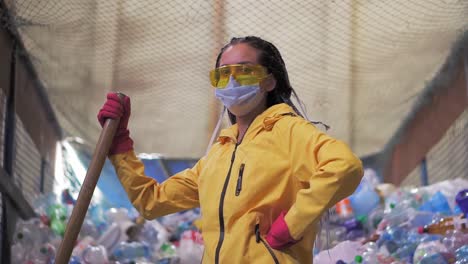 Portrait-Of-A-Girl-With-Dreadlocks,-In-Yellow-Jacket-And-Mask,-Holding-Large-Duck-Shovel,-Standing-At-A-Plastic-Recycling-Factory-1