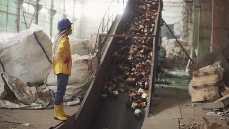 Woman-Worker-In-Yellow-And-Transparent-Protecting-Glasses,-Hard-Hat-And-Mask-Watching-The-Conveyor-Full-Of-Used-Plastic-Bottles-Lifting-Up