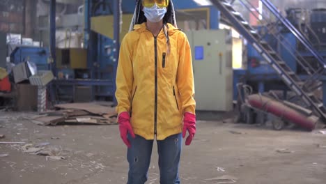 A-Young-Girl-With-Dreadlocks-In-Protective-Working-Clothes-Yellow-Jacket,-Eyeglasses,-Hard-Hat-And-Mask-Standing-Beyond-Recycling-Factory