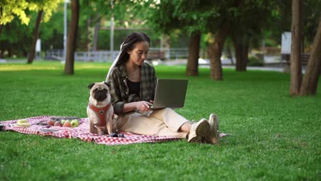 Young-Girl-Sitting-Outdoors-On-The-Grass-Lawn-With-Her-Laptop-Computer-And-Cute-Little-Pug-Is-Sitting-Near